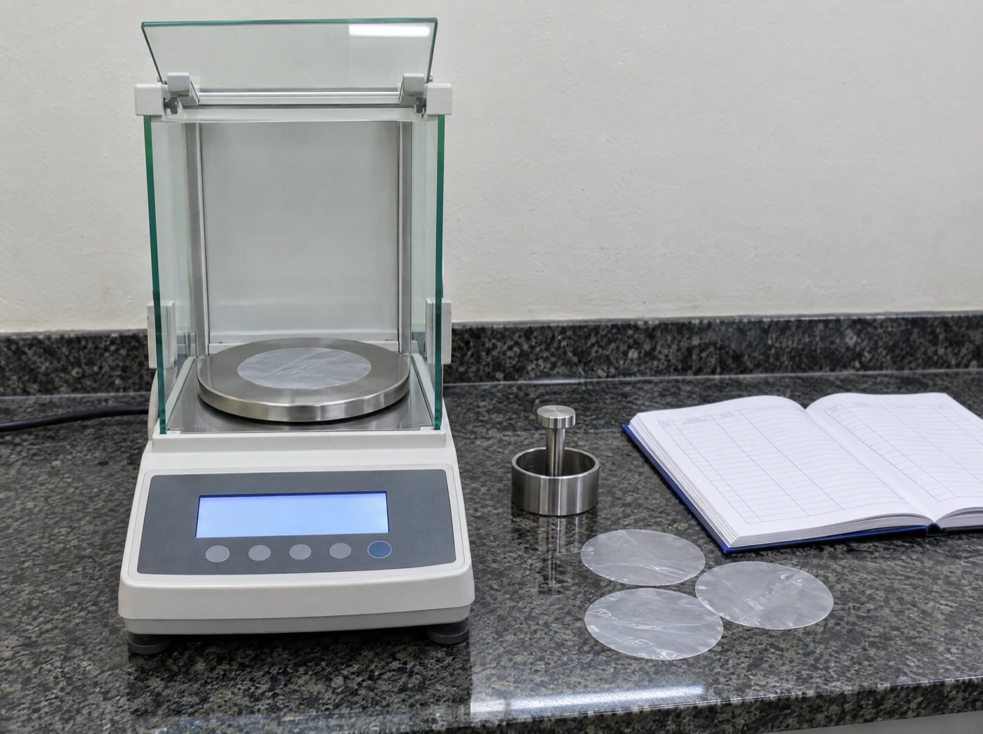Precision balance with a cut disc of film on the pan, sample cutter and record book beside it